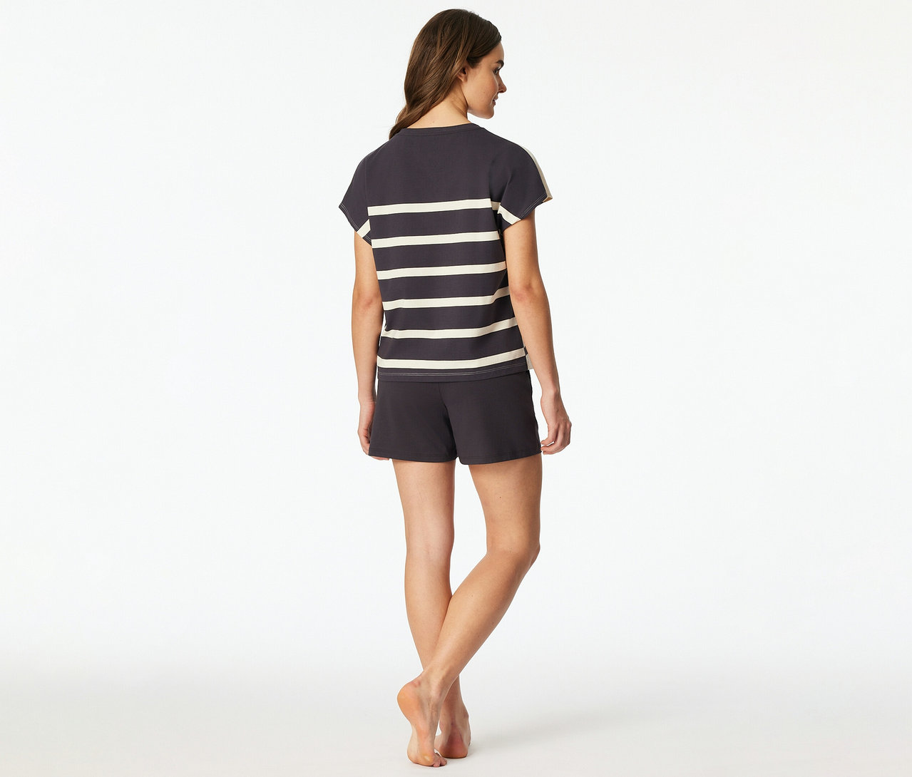 Une femme est debout, dos au spectateur, vêtue d'un set shorty SCHIESSER gris foncé avec un haut rayé et un short assorti. Elle est pieds nus.