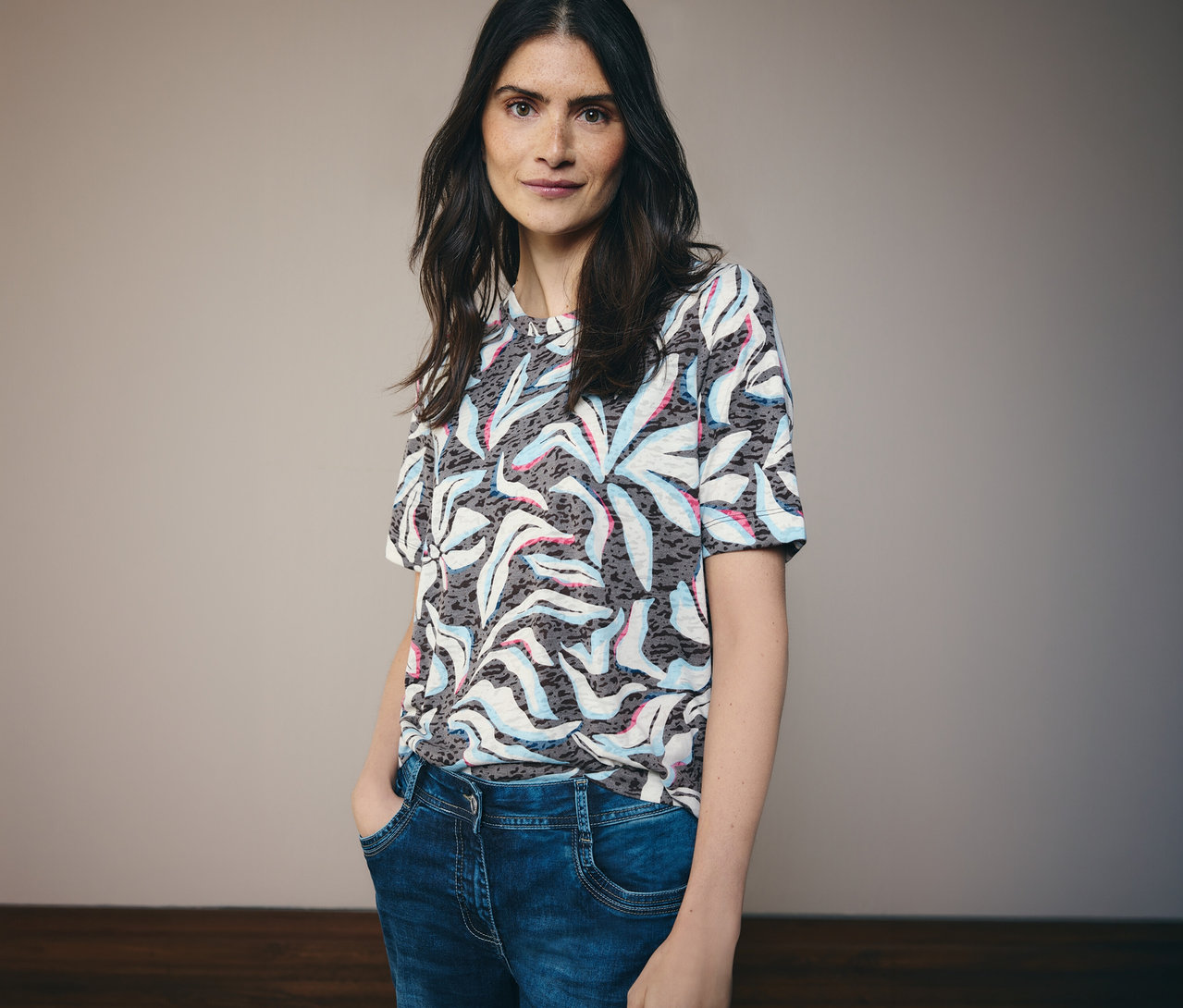 Une femme aux cheveux foncés porte un t-shirt manches courtes CECIL bleu à motifs et un jean bleu.