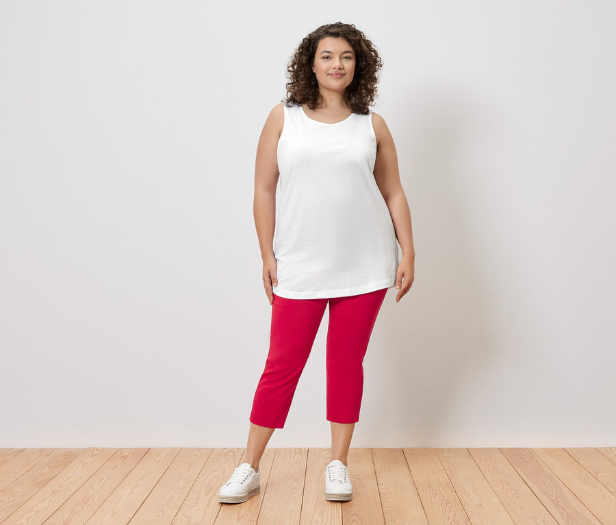 Une femme aux cheveux bouclés se tient devant un mur, vêtue d'un haut blanc, d'un pantalon rouge et de baskets blanches.