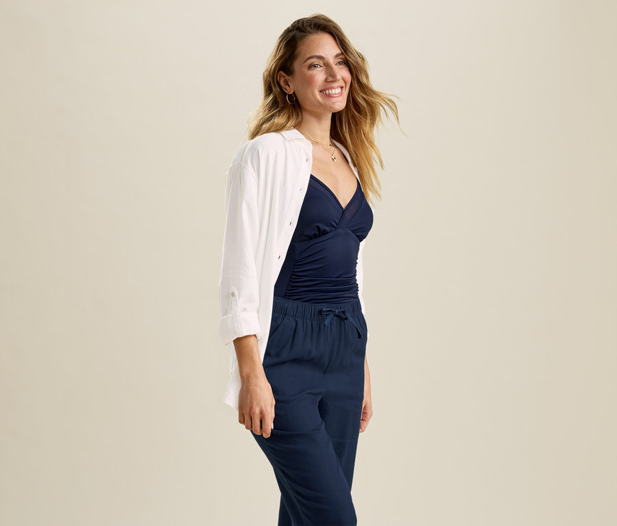 Femme souriante portant une blouse longue en lin blanche ouverte sur un haut bleu marine et un pantalon bleu marine.