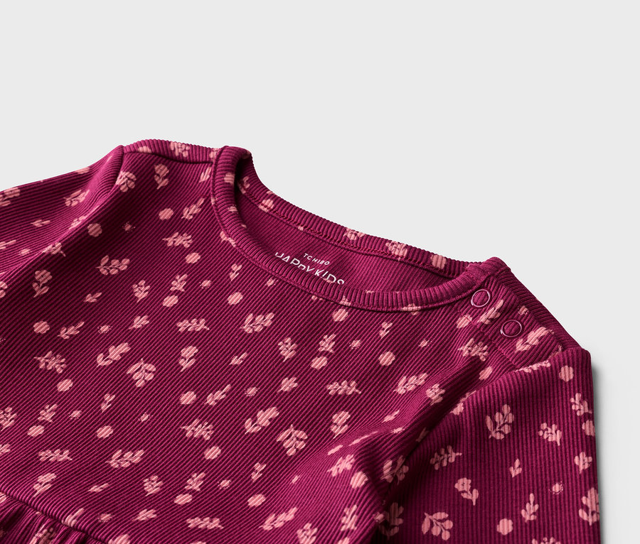 Gros plan sur un t-shirt bordeaux côtelé à manches longues avec un motif floral rose clair. Deux boutons-pression sont visibles sur l'épaule.