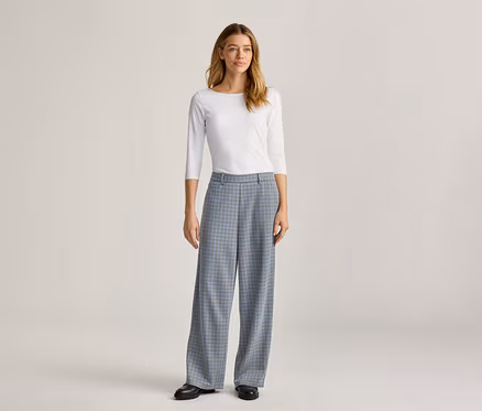 Une femme debout porte un haut blanc à manches longues et un pantalon large à carreaux bleus avec des chaussures noires.