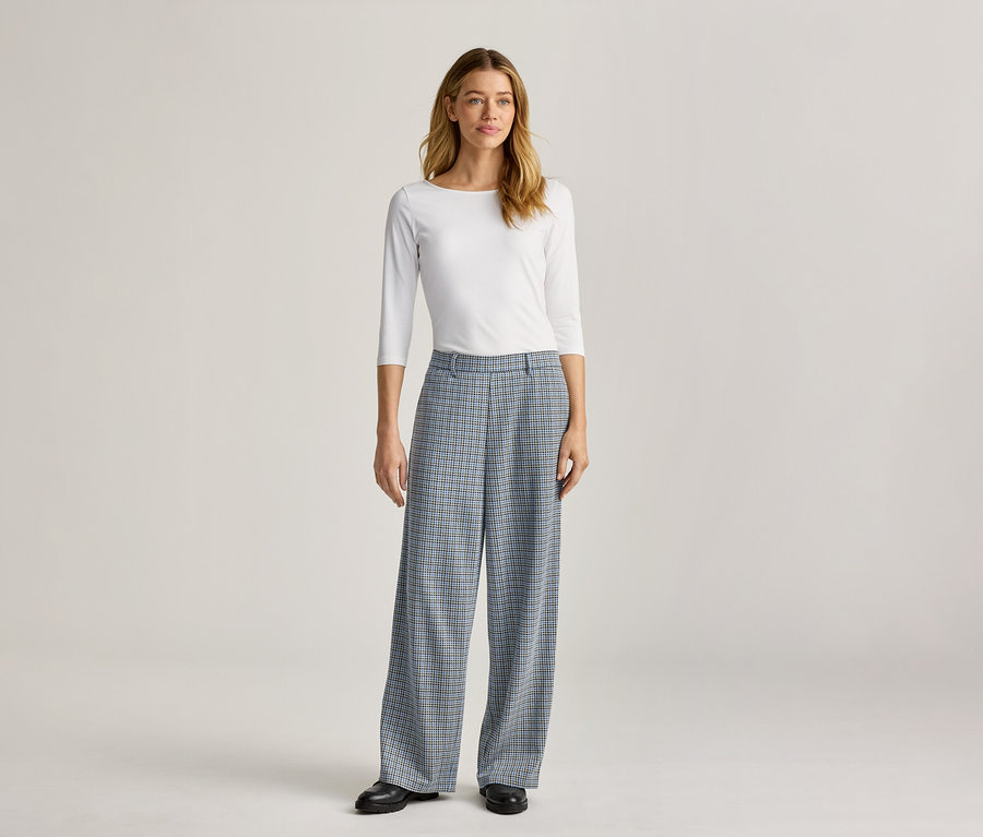 Une femme debout porte un haut blanc à manches longues et un pantalon large à carreaux bleus avec des chaussures noires.