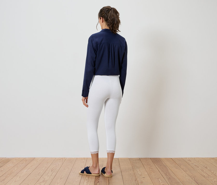 Une femme se tient dos à la caméra, vêtue d'un chemisier bleu marine, d'un legging blanc et de sandales bleues.