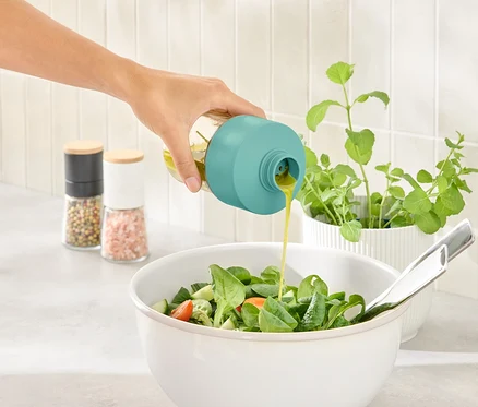 Une main verse de la vinaigrette d'une bouteille avec un bouchon turquoise dans un saladier.