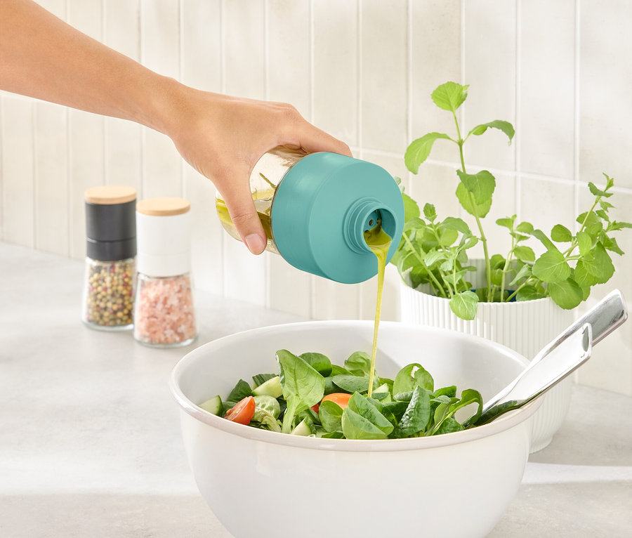 Une main verse de la vinaigrette d'une bouteille avec un bouchon turquoise dans un saladier.