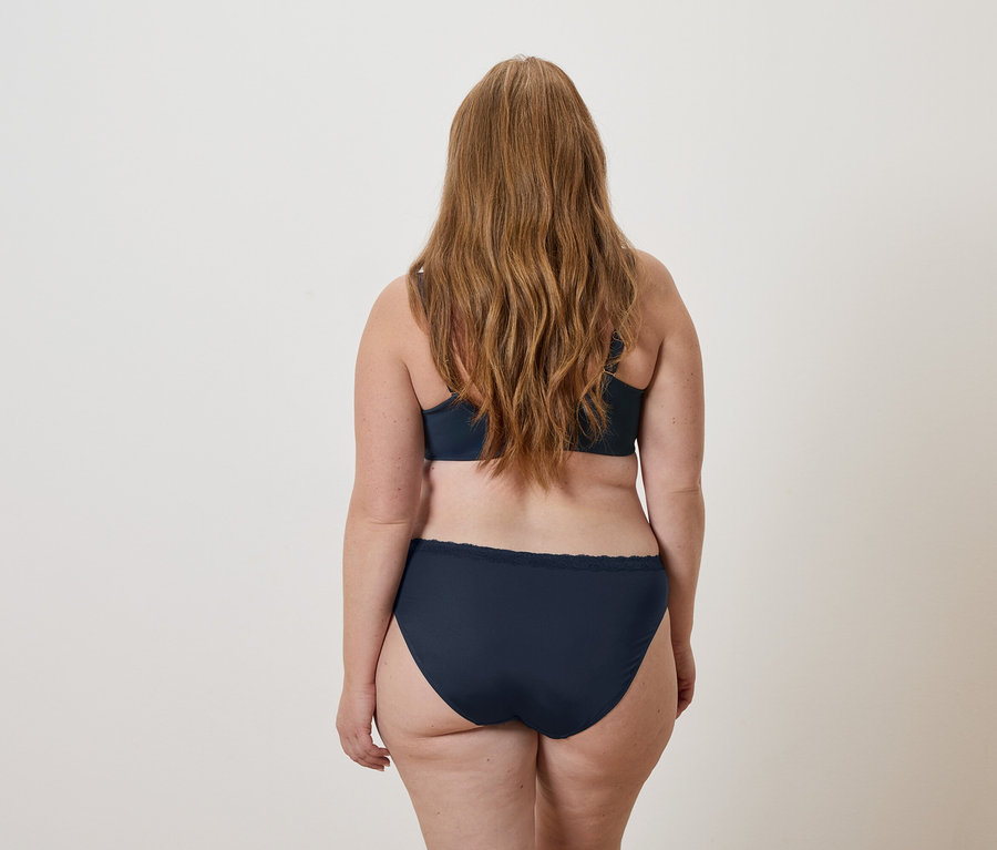 Vue arrière d'une femme aux longs cheveux ondulés blond roux portant un soutien-gorge bleu marine et des sous-vêtements assortis.