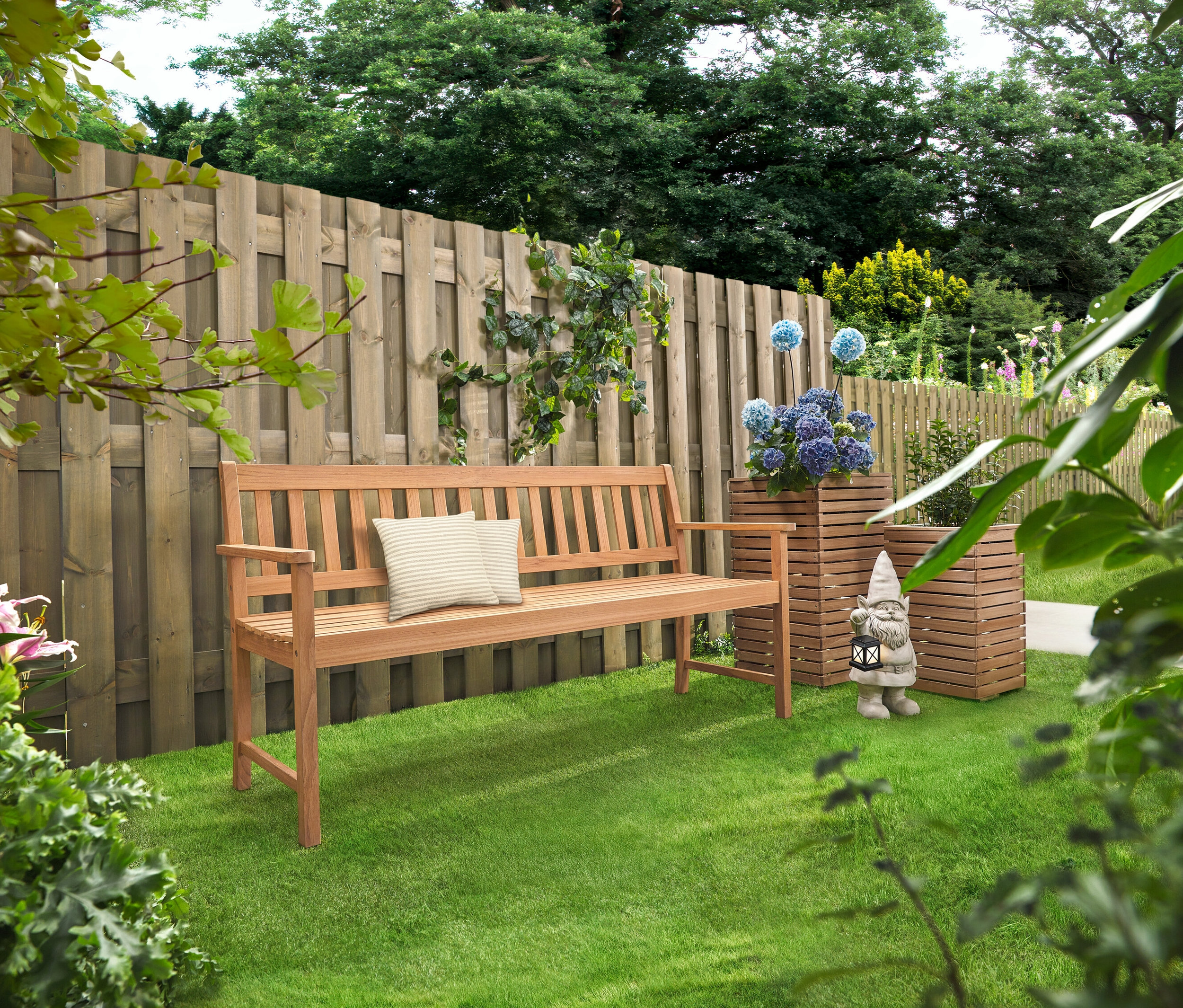 Un banc en bois avec des coussins se trouve sur une pelouse. Derrière se trouvent deux colonnes de plantation »Tinus« avec des fleurs bleues.