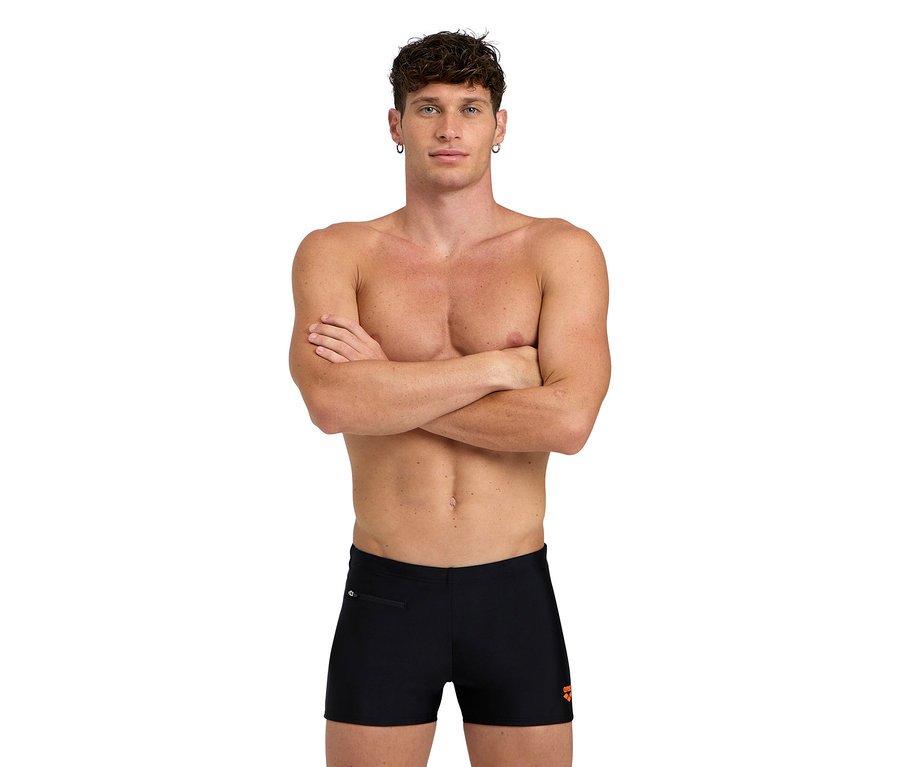 Homme les bras croisés, vêtu d'un short de bain noir.