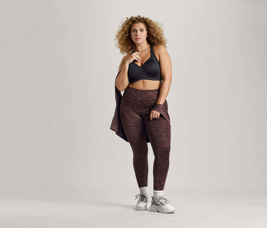 Femme aux cheveux bouclés portant un soutien-gorge de sport noir et un legging marron à motifs.