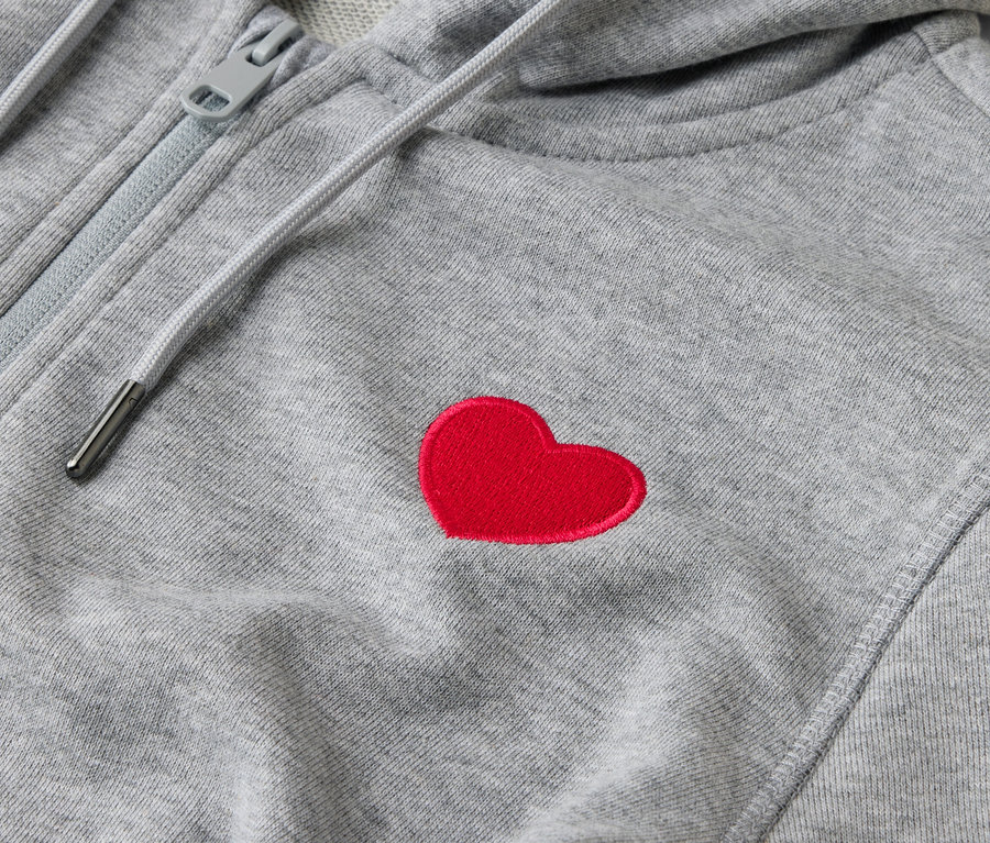 Gros plan sur un sweat à capuche gris zippé avec un patch en forme de cœur rouge.
