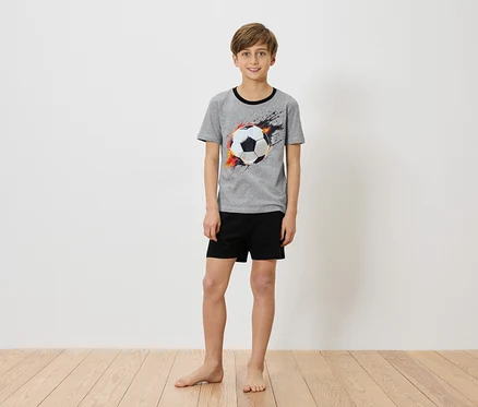 Un garçon se tient pieds nus devant un mur blanc, portant un t-shirt gris avec un imprimé de ballon de foot et un short noir.