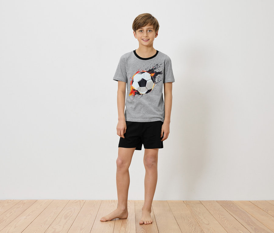 Un garçon se tient pieds nus devant un mur blanc, portant un t-shirt gris avec un imprimé de ballon de foot et un short noir.