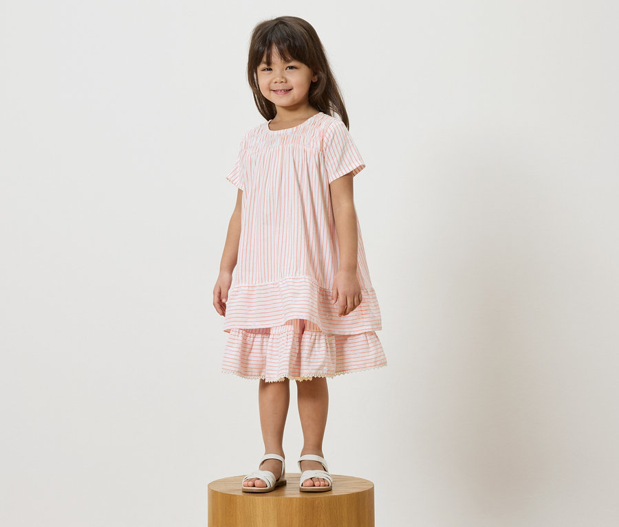 Fillette debout sur un tabouret portant une robe rayée rose et des sandales blanches.
