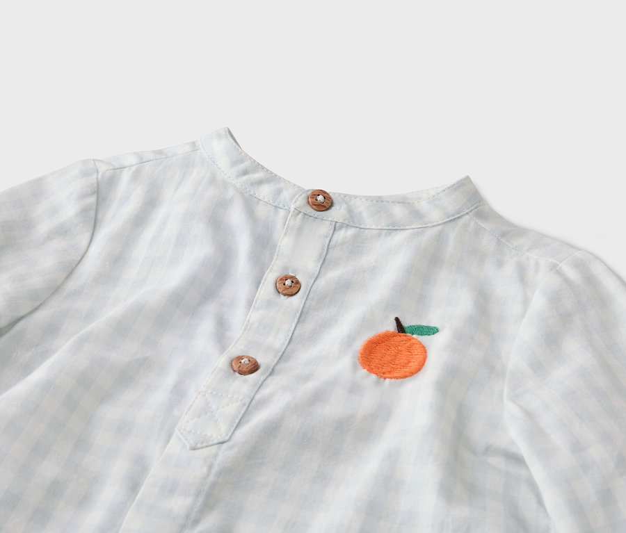 Gros plan d'une chemise à carreaux bleu clair et blanc avec un col mandarin, une patte de boutonnage à trois boutons et une orange brodée.