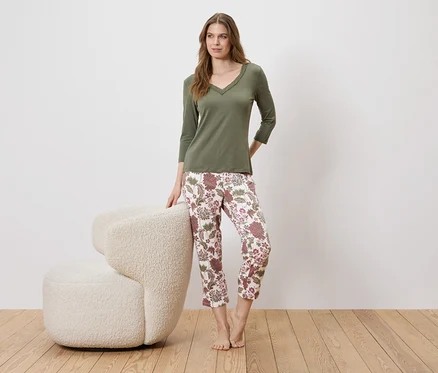 Une femme se tient à côté d'un fauteuil, portant un pull vert olive et un pantalon à fleurs.