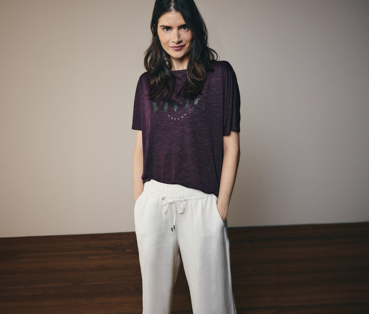 Une femme aux cheveux bruns ondulés portant un t-shirt bordeaux avec l'inscription « Vivid Dreams » et un pantalon blanc.