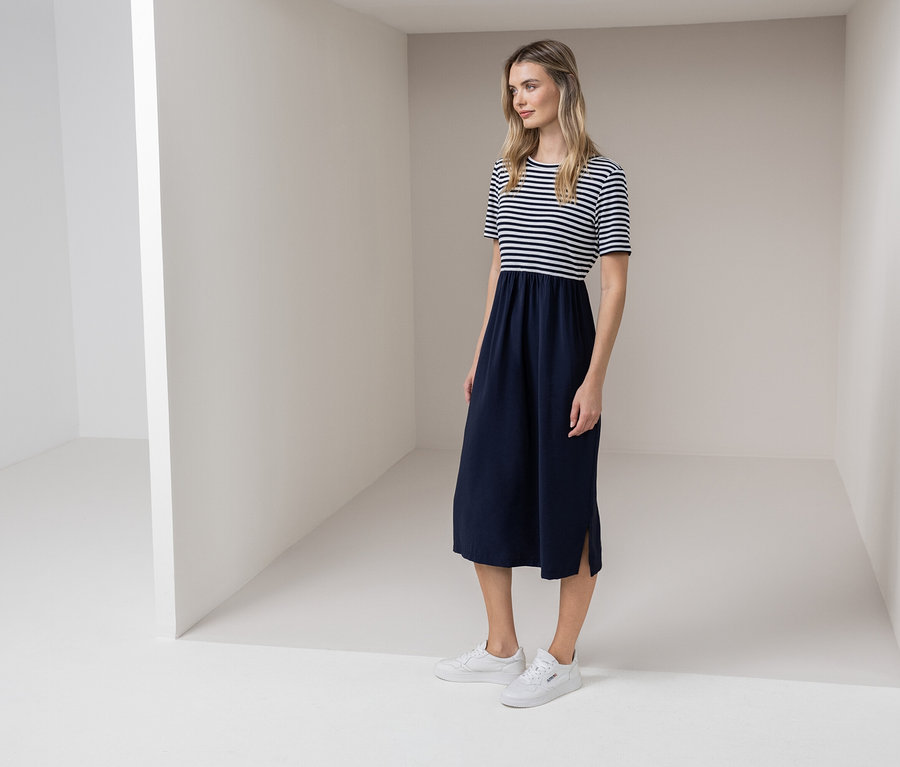 Une femme se tient dans une pièce blanche, portant une robe rayée bleu et blanc et des baskets blanches.
