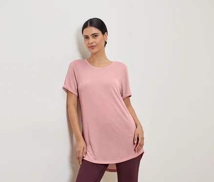 Une femme portant un t-shirt rose et des leggings, appuyée contre un mur blanc.