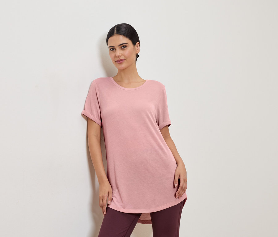 Une femme portant un t-shirt rose et des leggings, appuyée contre un mur blanc.