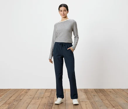 Femme portant un t-shirt à manches longues gris et blanc rayé et un pantalon bleu marine, les mains dans les poches, sur fond blanc.