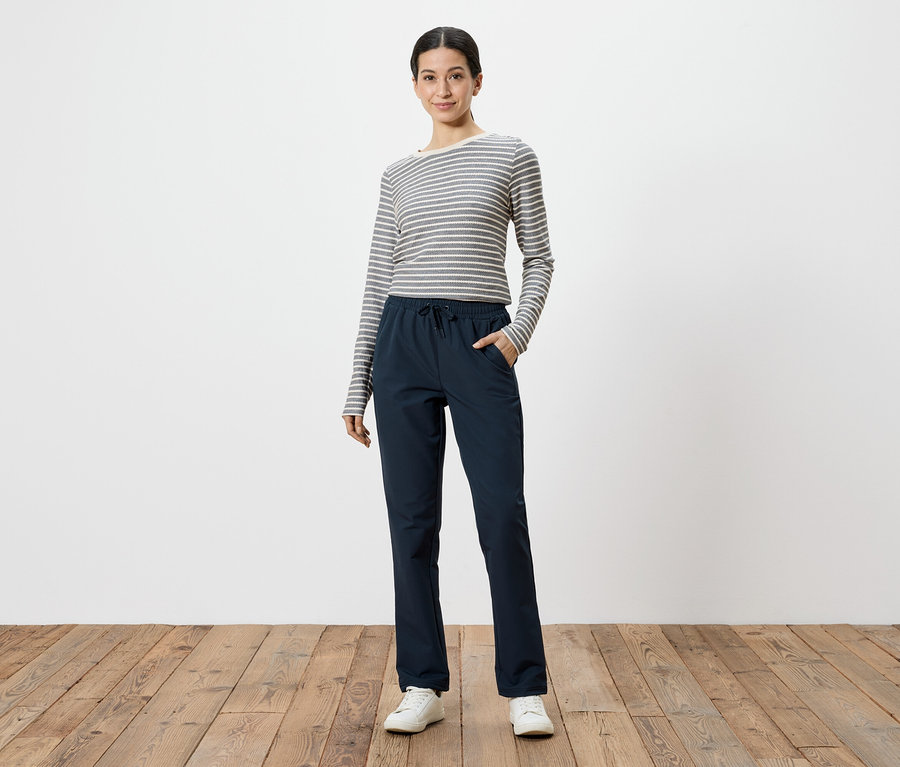 Femme portant un t-shirt à manches longues gris et blanc rayé et un pantalon bleu marine, les mains dans les poches, sur fond blanc.