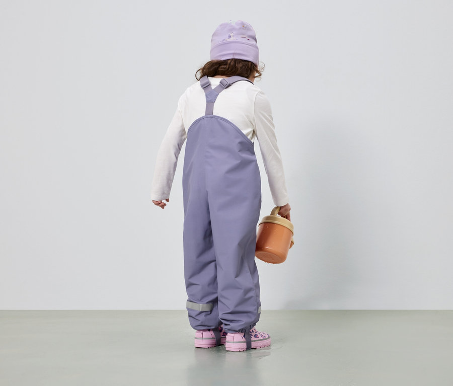 Vue arrière d'un enfant portant un bonnet et une salopette violets, un t-shirt blanc à manches longues, des chaussures roses et un seau orange.