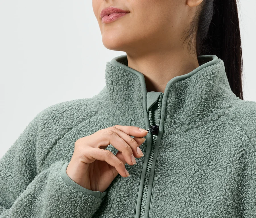 Une femme ferme un pull en polaire teddy vert clair. Le col et la fermeture éclair sont également vert clair.