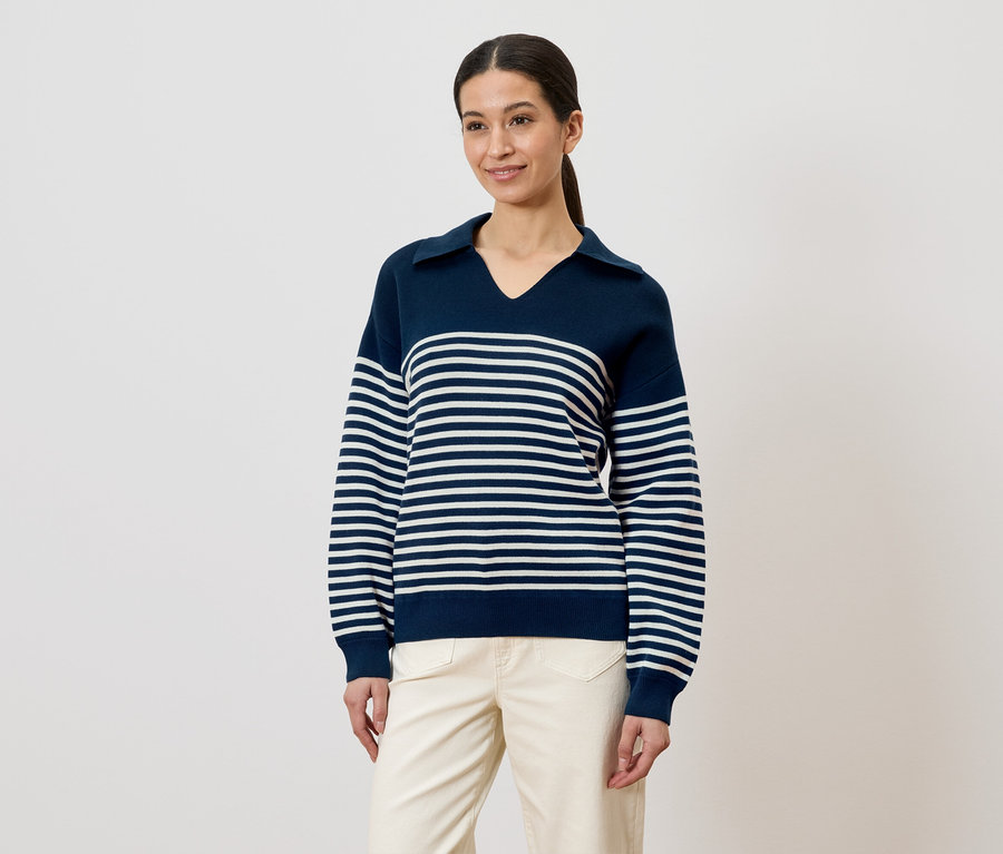 Une femme avec une queue de cheval porte un pull rayé bleu marine et un pantalon clair sur fond blanc.