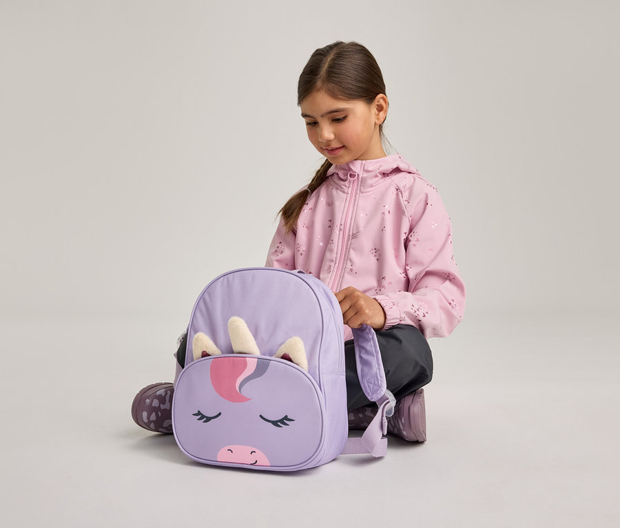 Une jeune fille en veste rose et pantalon foncé est accroupie sur le sol, regardant un sac à dos violet avec un motif de visage de licorne, incluant une corne et des oreilles.