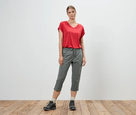 Une femme se tient devant un mur blanc, portant un t-shirt rouge et un pantacourt gris.