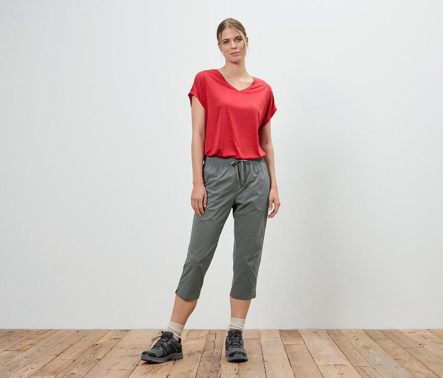 Une femme se tient devant un mur blanc, portant un t-shirt rouge et un pantacourt gris.