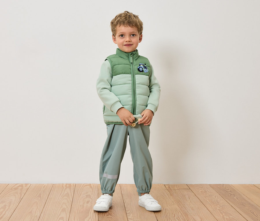 Un garçon se tient devant un mur, vêtu d'une veste vert clair, d'un pantalon vert clair et de baskets blanches.