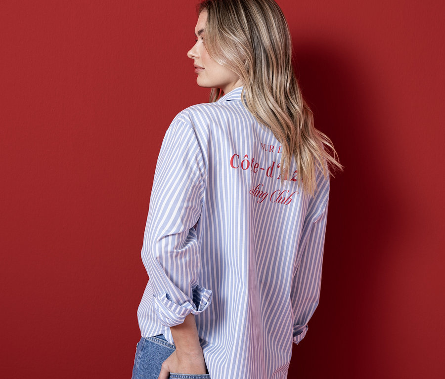 Une femme aux longs cheveux blonds, vue de dos et de profil, porte un chemisier Street One rayé bleu clair et blanc avec du texte rouge dans le dos, et un jean bleu. Sa main droite est dans sa poche arrière.
