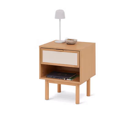 Table de chevet en bois clair avec un tiroir et une étagère ouverte. Une lampe blanche et un petit bol décoratif sont posés dessus, deux livres sont sur l'étagère.
