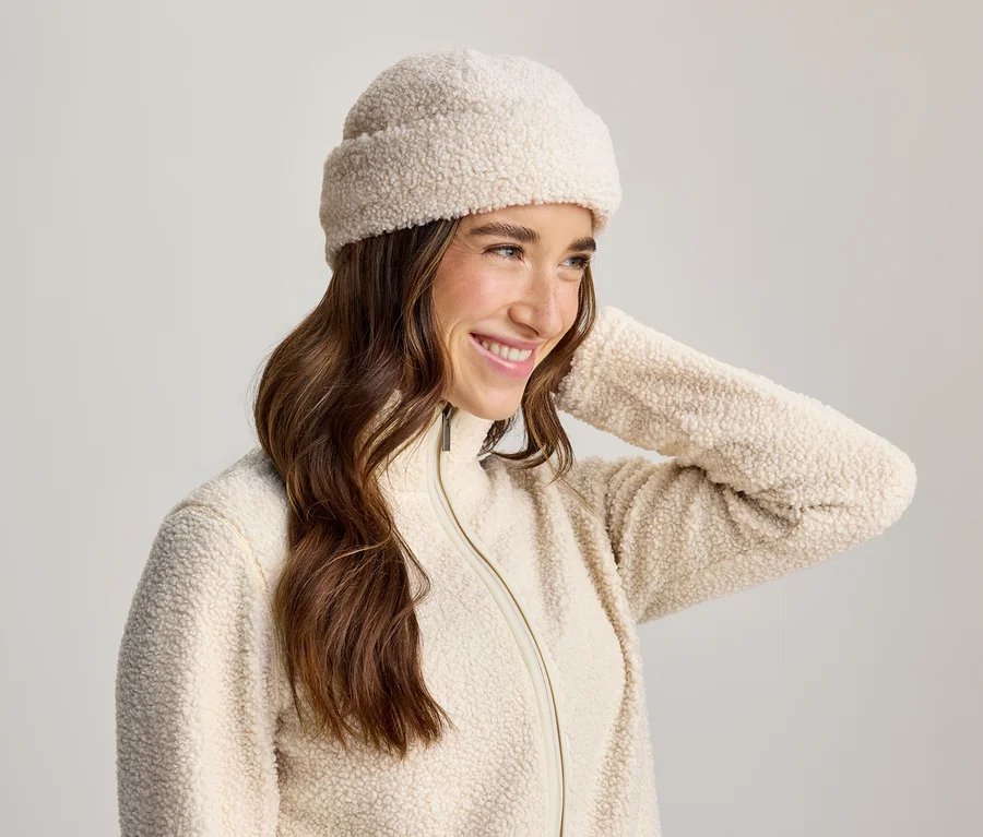Femme souriante aux longs cheveux bruns, portant un bonnet et une veste zippée en polaire couleur crème, la main derrière la tête.