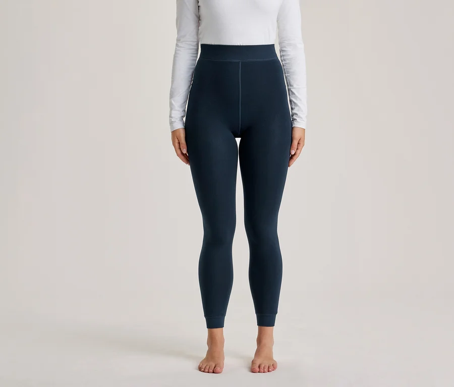 Personne portant un haut blanc à manches longues et un legging bleu foncé, debout pieds nus devant un fond uni.
