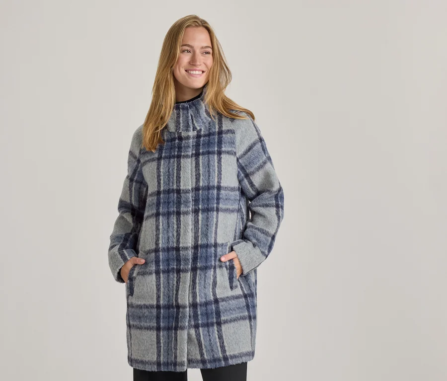 Femme souriante portant un manteau à carreaux gris et bleu avec un col montant, les mains dans les poches.