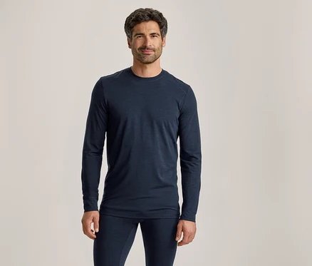 Un homme aux cheveux foncés et barbu, portant un haut à manches longues bleu foncé et un pantalon assorti, se tient debout et regarde devant lui.