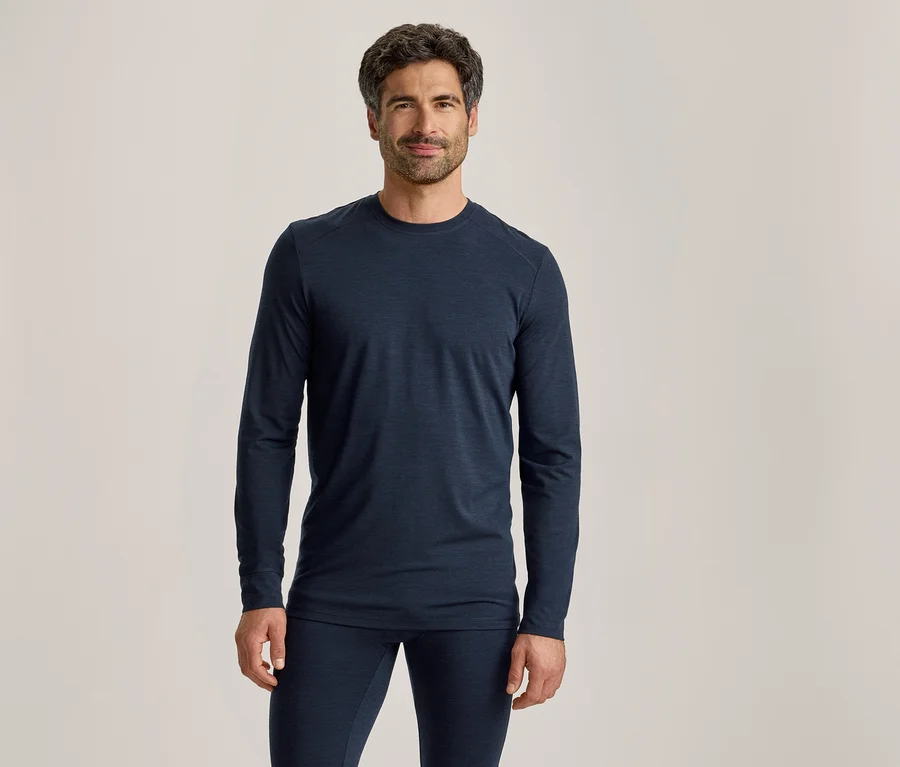 Un homme aux cheveux foncés et barbu, portant un haut à manches longues bleu foncé et un pantalon assorti, se tient debout et regarde devant lui.