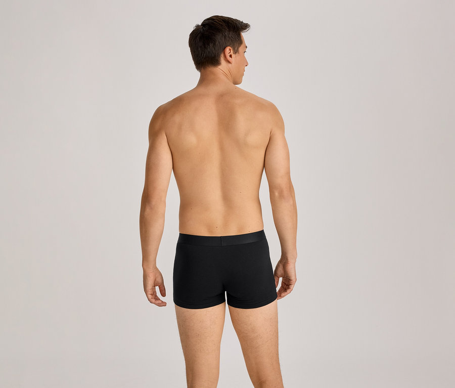 Un homme torse nu vu de dos, portant un boxer noir, debout devant un fond clair.