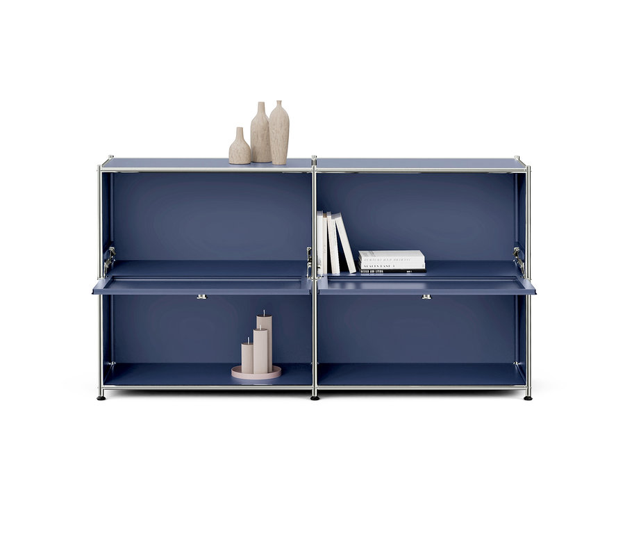 Une armoire modulaire bleue avec un cadre chromé, comprenant quatre compartiments. Trois vases beiges sont posés dessus. Le compartiment supérieur droit contient trois livres blancs, tandis que le compartiment inférieur gauche présente trois bougies beiges sur un plateau rose, avec sa porte légèrement ouverte. La porte inférieure droite est également légèrement ouverte.