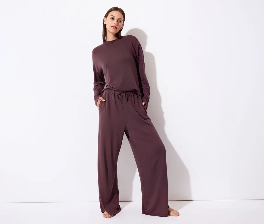 Femme debout, vêtue d'un haut à manches longues prune foncé et d'un pantalon large assorti, les mains dans les poches. Ses pieds nus sont visibles sur un fond blanc.