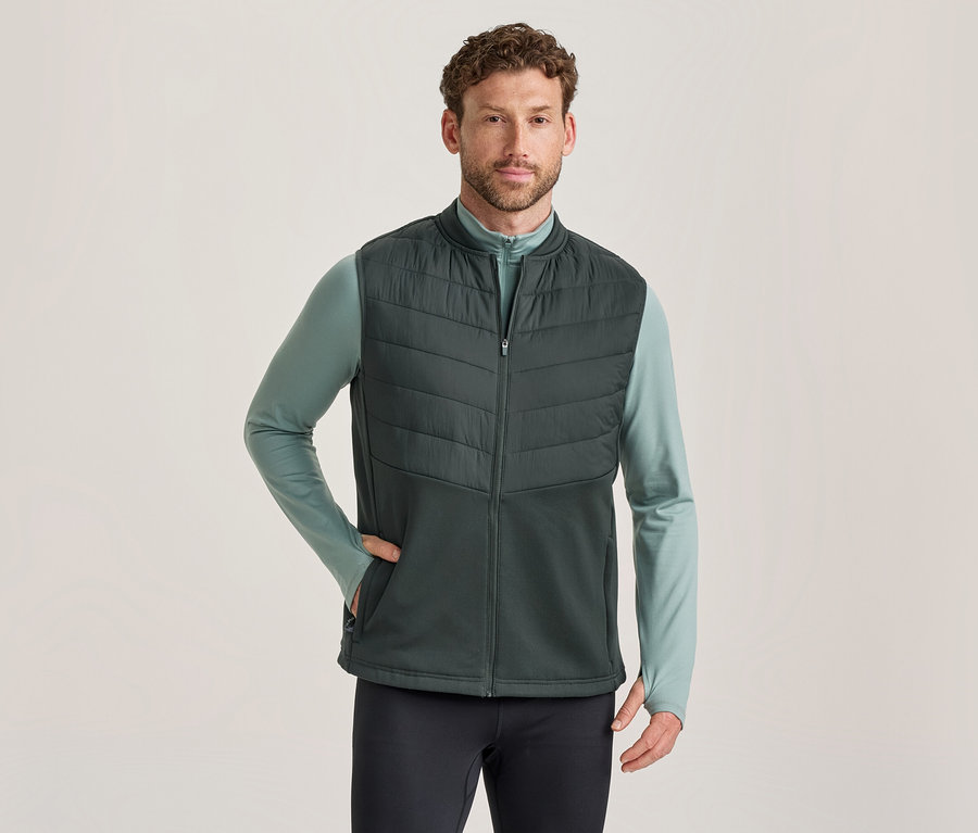 Homme portant un gilet matelassé vert foncé sur un t-shirt à manches longues vert clair et un pantalon foncé, une main dans la poche du gilet.