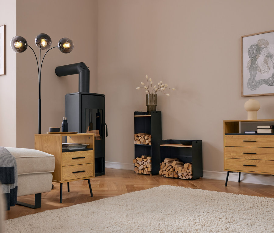 Salon moderne avec un poêle à bois noir, une étagère haute pour bois de chauffage avec surface de rangement et une étagère large pour bois de chauffage avec surface de rangement, toutes deux remplies de bois. Un canapé clair, une table en bois et une commode sont sur un tapis.