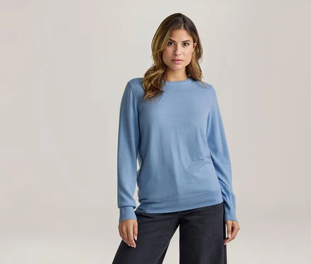 Jeune femme aux cheveux bruns portant un pull à manches longues bleu clair et un pantalon foncé, debout et regardant devant elle.