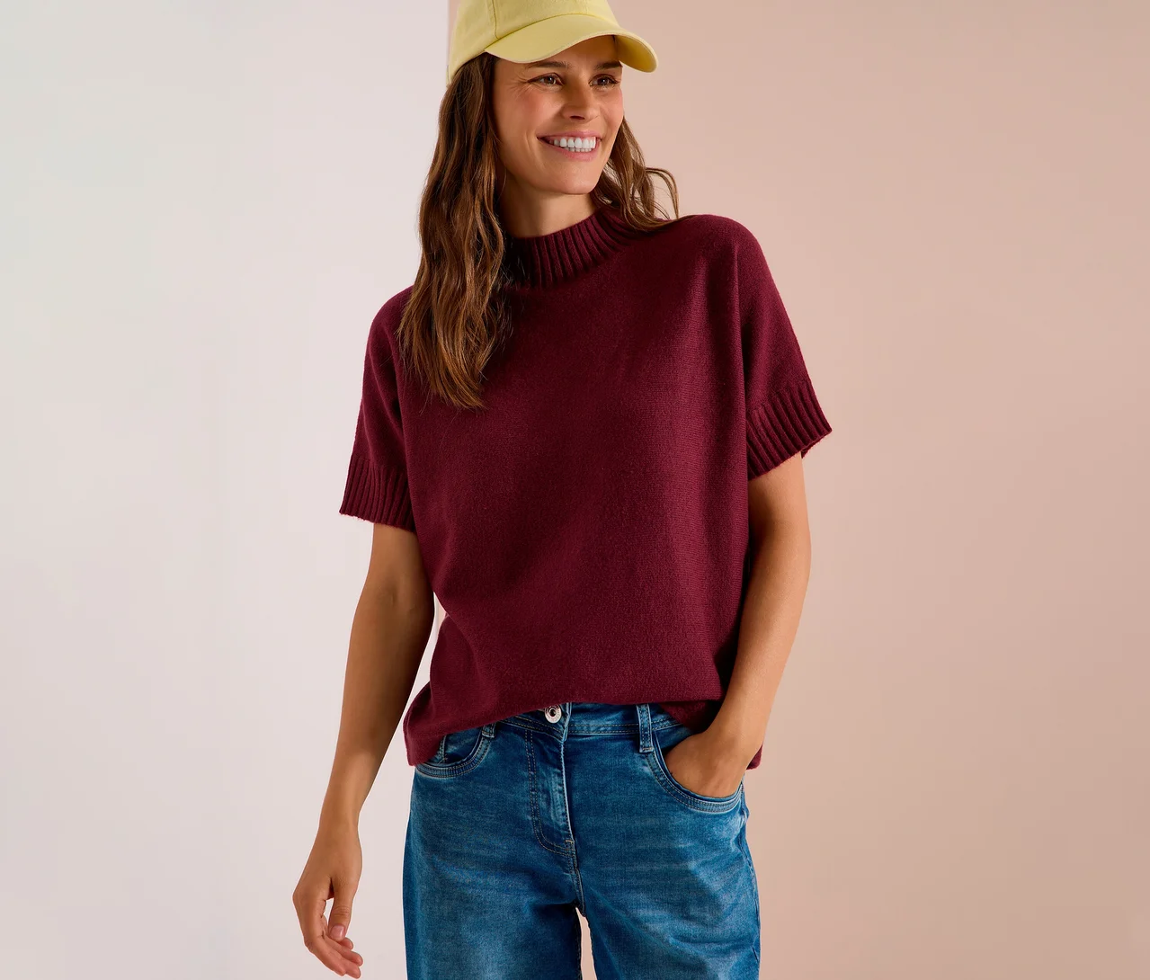 Femme souriante portant une casquette jaune, un pull en maille CECIL rouge bordeaux et un jean.