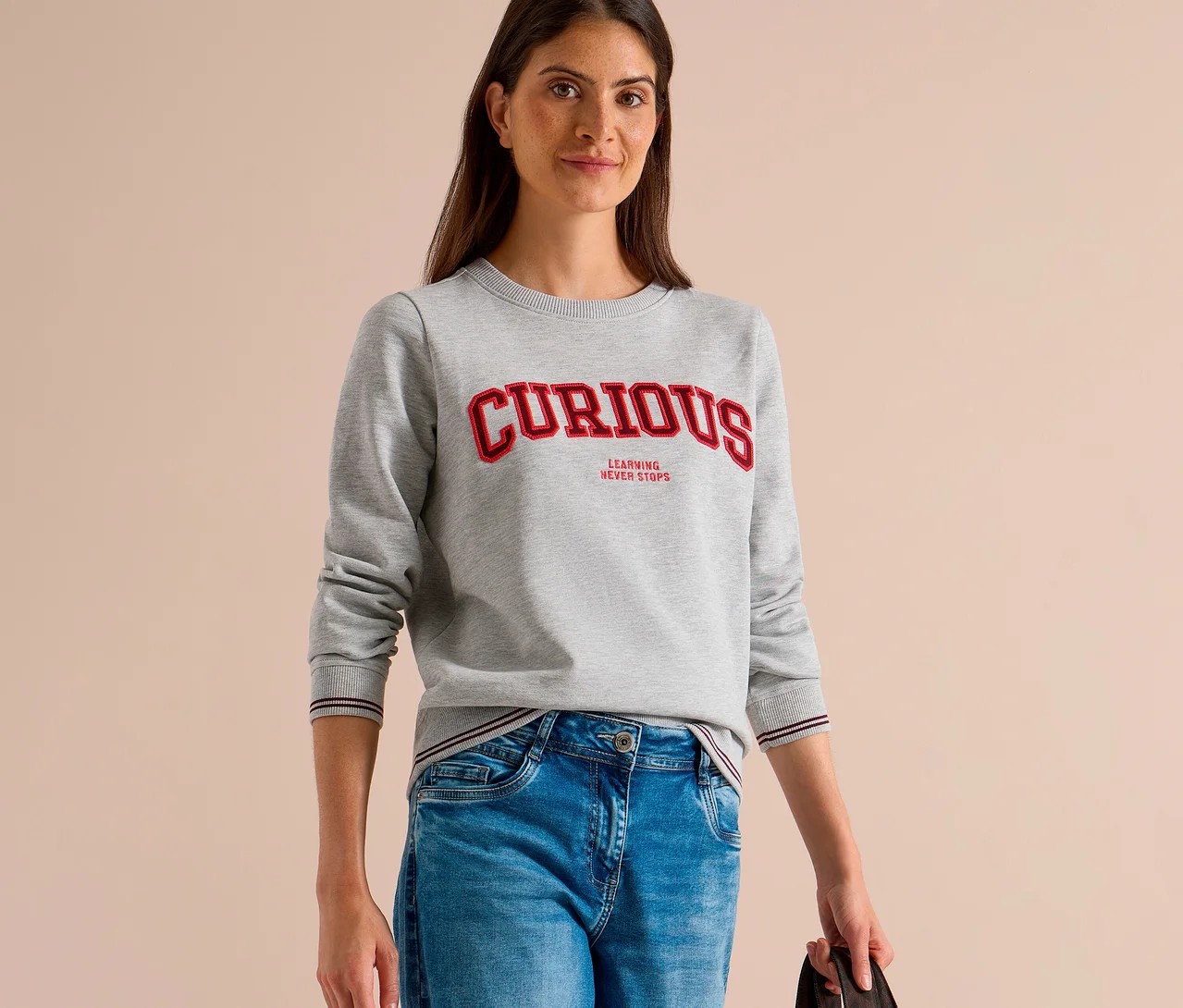 Une femme porte un sweatshirt CECIL gris avec l'inscription „CURIOUS LEARNING NEVER STOPS“ et un jean bleu.