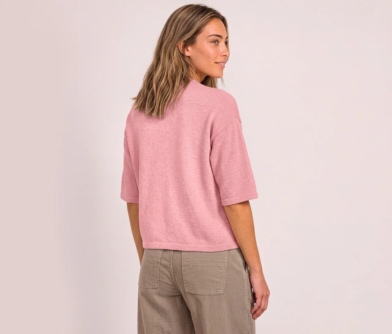 Une femme aux longs cheveux châtains clairs, vue de dos, regarde par-dessus son épaule droite. Elle porte un pull en maille 3/4 Street One rose et un pantalon en velours côtelé marron clair.