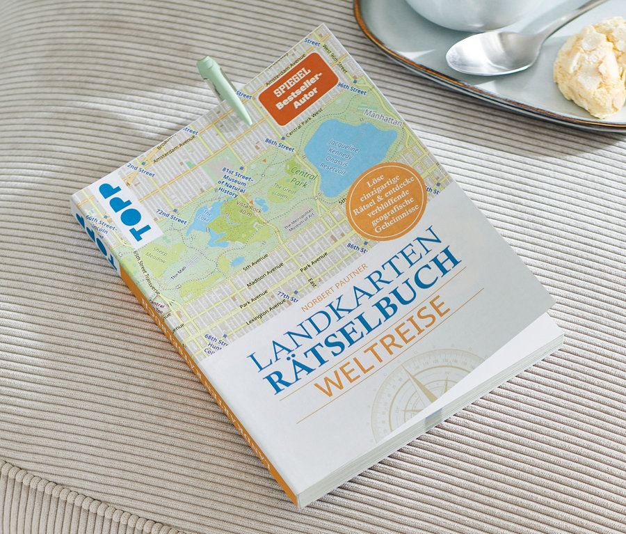 Sur un tabouret beige se trouve un livre : Landkarten-Rätselbuch « Weltreise », à côté d’une tasse de café et des biscuits.
